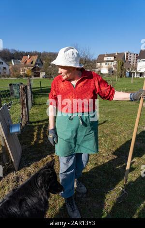 Gärtnerin sieht seitlich aus mit rotem Hemd, blauer Hose, grüner Schürze, weißem Hut und schwarzem Hund. Trait, rotem Hemd, blauer Hose, grüner Schürze, WH Stockfoto