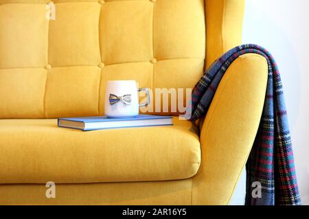 Tasse Tee und blaues Buch auf gelbem Trainer mit Decke. Details zum Stillleben im Inneren des Wohnzimmers. Gemütliches Innenleben, Hauskomfort Stockfoto