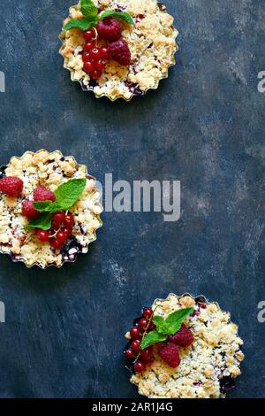 Portionsbeere zerbröckelt in einer Backform auf dunklem Hintergrund. Beerentartlets mit Mürbeteig und saftiger Füllung. Leichte Sommerdessert. Stockfoto