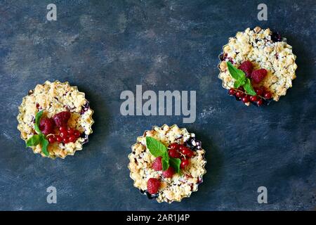 Portionsbeere zerbröckelt in einer Backform auf dunklem Hintergrund. Beerentartlets mit Mürbeteig und saftiger Füllung. Leichte Sommerdessert. Stockfoto
