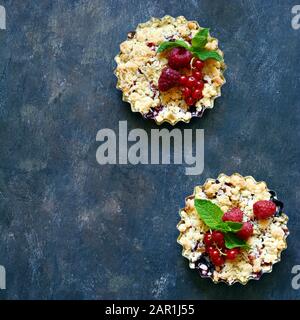 Portionsbeere zerbröckelt in einer Backform auf dunklem Hintergrund. Beerentartlets mit Mürbeteig und saftiger Füllung. Leichte Sommerdessert. Stockfoto