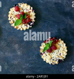 Portionsbeere zerbröckelt in einer Backform auf dunklem Hintergrund. Beerentartlets mit Mürbeteig und saftiger Füllung. Leichte Sommerdessert. Stockfoto