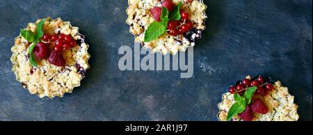 Portionsbeere zerbröckelt in einer Backform auf dunklem Hintergrund. Beerentartlets mit Mürbeteig und saftiger Füllung. Leichte Sommerdessert. Stockfoto