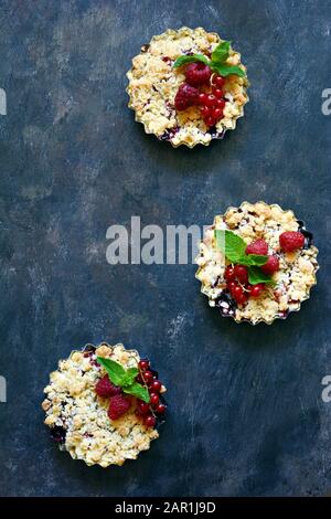 Portionsbeere zerbröckelt in einer Backform auf dunklem Hintergrund. Beerentartlets mit Mürbeteig und saftiger Füllung. Leichte Sommerdessert. Stockfoto