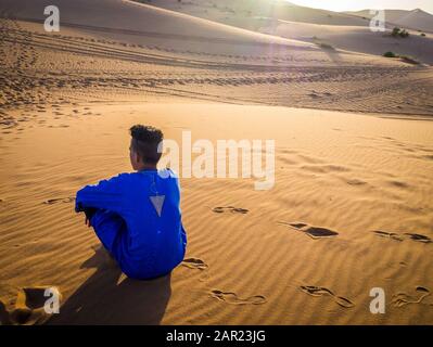 Merzouga, MAROKKO - 07. Juni 2018: Beduinenjunge mit blauem Djellaba und orangefarbenem Schal in der Wüste Stockfoto