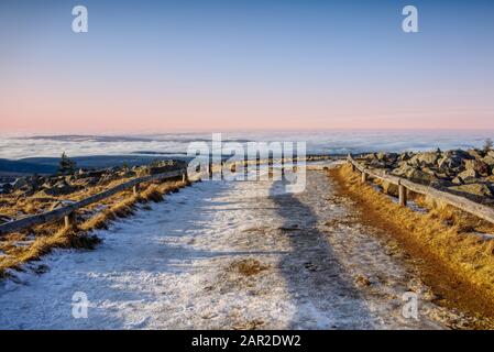 Schneebedeckter Wanderweg über einem Wolkenmeer und einem Himmel in Pastelle. Romantische Winterlandschaft auf dem Brocken, Harz, Deutschland. Stockfoto