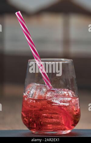 Nahaufnahme eines leckeren Glases mit rotem Getränk mit Eiswürfeln und einem Strohhalm auf einem unscharf wirkenden Hintergrund. Freizeit- und Entspannungskonzept. Stockfoto