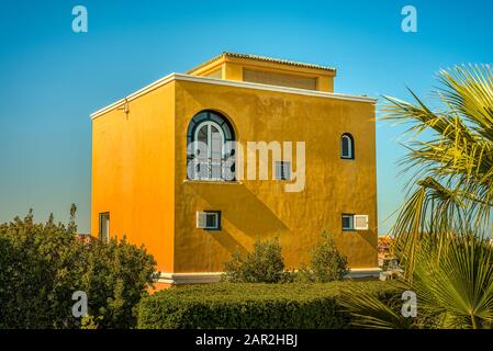 Ein quadratisches gelbes Haus unter den Palmen im Sonnenschein gegen einen blauen Himmel, El Gouna, Ägypten, 12. Januar 2020 Stockfoto