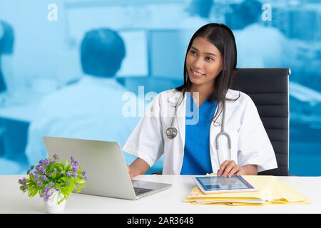 Weibliche Ärztin lächelte willkommen, während sie an einem Notebook und einem Computertablett arbeitete. Stockfoto