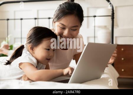 Fröhliche asiatische Mutter und kleine Tochter sehen sich Videos auf dem Laptop an Stockfoto