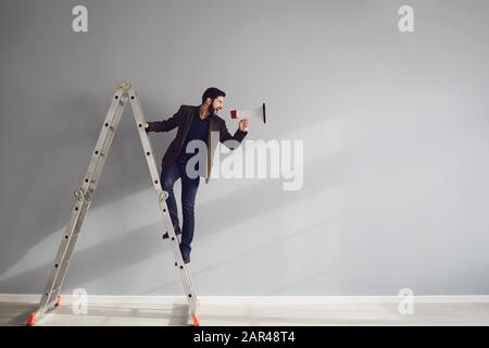 Ein bärtiger Geschäftsmann auf der Treppe mit einem Megafon in der Hand schreit auf grauem Hintergrund. Stockfoto