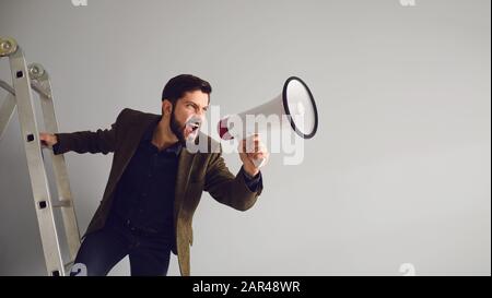 Ein bärtiger Geschäftsmann auf der Treppe mit einem Megafon in der Hand schreit auf grauem Hintergrund. Stockfoto