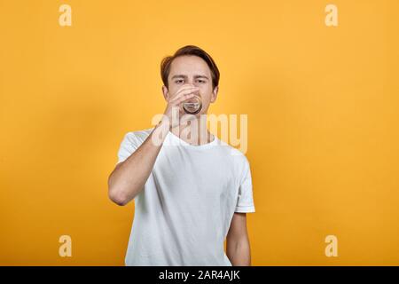 Der Mann trinkt Wasser aus einem Glas, in weißem T-Shirt gelbem Hintergrund. Stockfoto