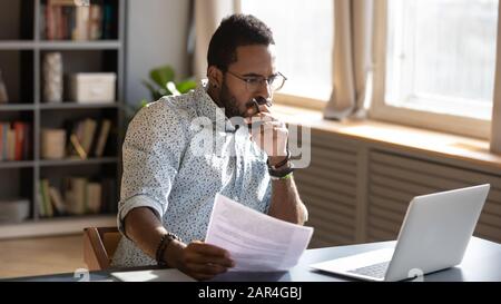 Ein fokussierter afrikanischer Geschäftsmann, der Dokumente mit Blick auf einen Laptop hält, der recherchiert Stockfoto