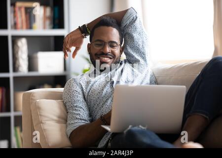 Lächelnder junger afrikanischer Erwachsener, der auf dem Sofa einen Laptop zum Entspannen verwendet Stockfoto