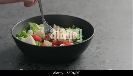 Man handpflücken Salat mit Mozzarella, Kirschtomaten und Friseeblättern in schwarzer Schüssel auf Terrazzooberfläche, breites Foto Stockfoto