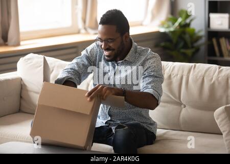 Lächelnder afrikanischer Mann, der ein Kartonpaket auf dem Sofa öffnet Stockfoto