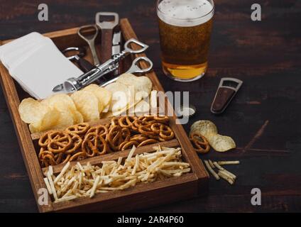 Glas Handwerk Lagerbier und Opener mit Box von Snacks auf dunklen Hintergrund. Brezeln und Chips und salzig Kartoffelstifte in vintage Holzbox mit Stockfoto