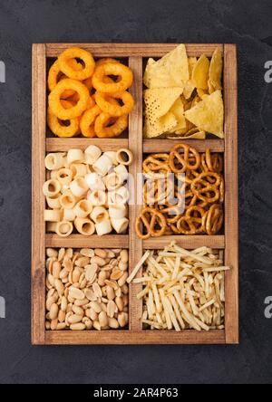 Verschiedene Snacks in Vintage hölzerne Kiste auf schwarze Küche Hintergrund. Zwiebelringe, Nachos, salzige Erdnüsse mit Potato Sticks und Brezel. Geeignet für Bier Stockfoto