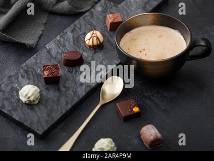 Luxus Pralinen Auswahl mit Tasse Cappuccino Kaffee und Dessert Löffel auf schwarzem Marmor und dunklem Hintergrund. Stockfoto