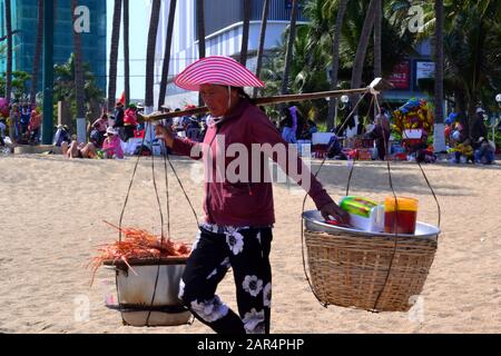 Ein Lebensmittelverkäufer von Frauen trägt zwei Körbe an einem Schulterpol entlang des Strandes in Nha Trang in Vietnam, Asien, Südostasien, an einem heißen, sonnigen Tag während des Tet-Neujahrs. Stockfoto