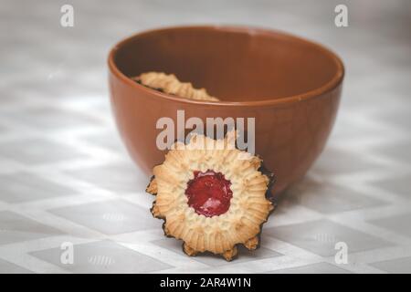 Knusprige Teigkekse mit Erdbeermarmelade und einem schokoladenbeschichteten Boden. In einer roten Töpferschüssel. Vor einem verschwommenen weißen Hintergrund. Stockfoto