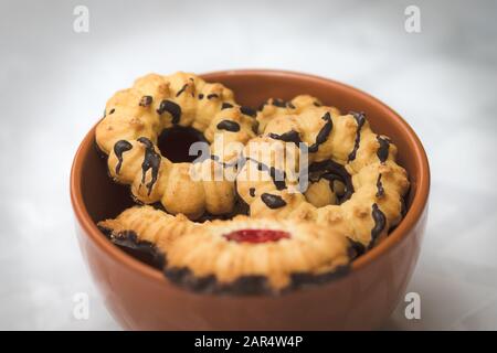 Knusprige Teigkekse mit Erdbeermarmelade und einem schokoladenbeschichteten Boden. In einer roten Töpferschüssel. Vor einem verschwommenen weißen Hintergrund. Stockfoto