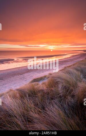 Ein lebendiger, leuchtend orangefarbener Sonnenaufgang von den Sanddünen über dem Strand der Druridge Bay und der Nordsee an der Northumberland-Küste im Winter.