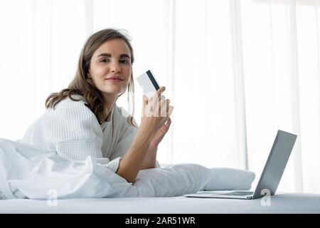 Schöne junge kaukasische Frau in weißem Pyjama, die online mit einem Laptop einkauft und mit einer Kreditkarte für Waren bezahlt, die auf einem weißen Bett im Schlafzimmer bestellt wurden Stockfoto