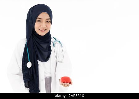 Asiatischer junger Erwachsener attraktiver muslimischer Frauenarzt, der mit Stethoskop stehend Hijab trägt und rotes Herz in weißem Hintergrund isoliert hält. Healthcar Stockfoto