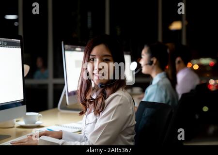 Asiatische junge Erwachsene vertrauen Betreiber Frau Agent mit Headsets, die in einem Call Center bei Nacht Umwelt mit seinem Kollegen Team als Kunde serv Stockfoto