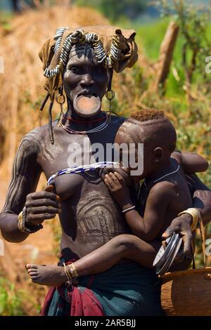 Mursi Frau stillt Baby und verkauft Teller. Die Mursi-Frauen sind berühmt dafür, Platten in ihren unteren Lippen zu tragen. Der Grund für diese "Verzierung" ist Stockfoto