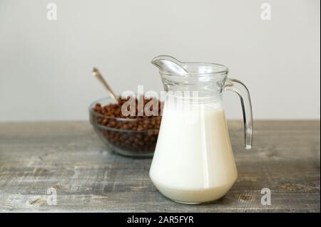 Auf dem Tisch befindet sich eine transparente Kanne mit frischer Milch und im Hintergrund eine Tasse mit Schokoladenflocken in Form von Bällen. Schnelles Frühstück im ho Stockfoto