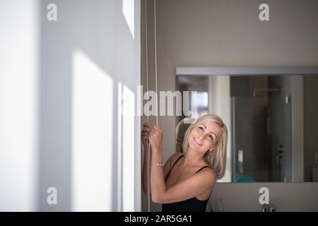 Hübsche, junge Frau, die Senkung der Innenbereich Jalousien/Rollos in ihrer modernen Interieur Wohnung Stockfoto