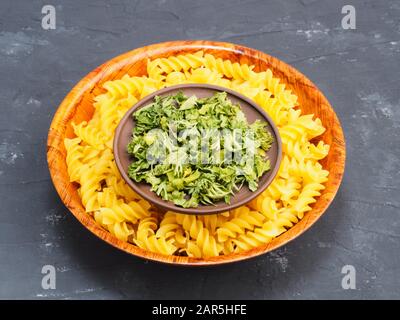 Pasta in Holzschale und trockenes grünes Gras in Tonplatte auf schwarzem Betongrund. Gesundes Esskonzept Stockfoto