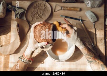 Craftsperson Konzept. Junge Frau, die in Innenräumen Keramik anstellt, entfernt Staub mit Schwamm, Oberansicht, Abschluss Stockfoto