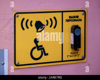 Ein Schild an der rosafarbenen Wand des Gebäudes mit einem Rollstuhllogo und einer Anruftaste für das Personal. Gelbe rechteckige Platte mit Klingel und Gegensprechanlage. Leer f Stockfoto