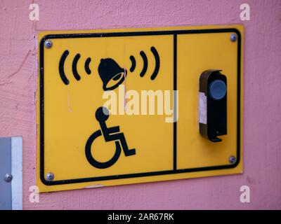 Ein Schild an der rosafarbenen Wand des Gebäudes mit einem Rollstuhllogo und einer Anruftaste für das Personal. Gelbe rechteckige Platte mit Klingel und Gegensprechanlage. Leer f Stockfoto