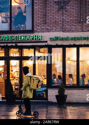 Moskau, Russland - 17. Januar 2020: Restaurantfenster am Abend. Die Leute sitzen an Tischen. Yandex Eda-Kurier fährt mit einem gelben Thermalbad vorbei Stockfoto