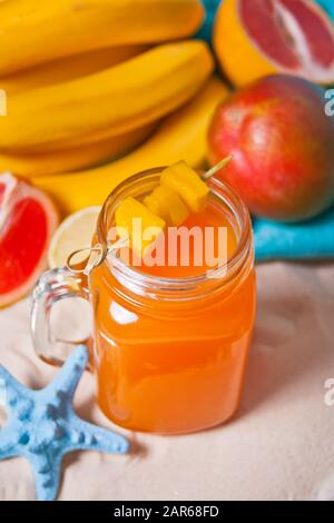 Glas tropischer exotischer Multifruchtsaft auf dem Sand mit Früchten im Hintergrund. Picknick am tropischen Strand. Stockfoto