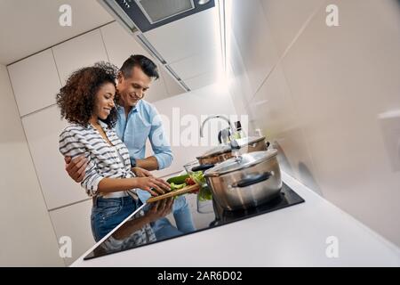 Romantisches Datum. Junges multiethnisches Paar, das in der Küche kocht, zum Abendessen Gemüse für Salat zuschneiden, fröhlich Stockfoto