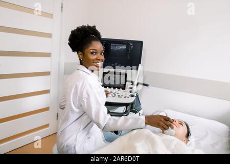 Lächelnder afroamerikanischer Frauenarzt, der den Ultraschall der Schilddrüse für weibliche Patienten macht. Medizinische Untersuchung einer jungen kaukasischen Frau durch Ultraschall Stockfoto