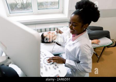 Attraktive junge Frau, die einen Schilddrüsen-Ultraschalltest von ihrem afrikanischen Arzt in der modernen Klinik durchführen kann Stockfoto