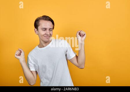 Der junge Mann in einem weißen T-Shirt auf gelbem Hintergrund hebt seine Hände im Glück. Stockfoto