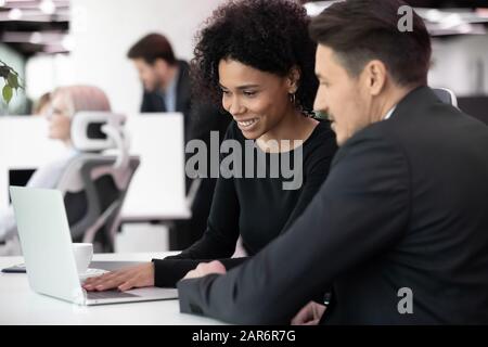 Zufriedene Kollegen aus verschiedenen Rassen, die das Projekt am Arbeitsplatz auf einem Laptop verwalten. Stockfoto