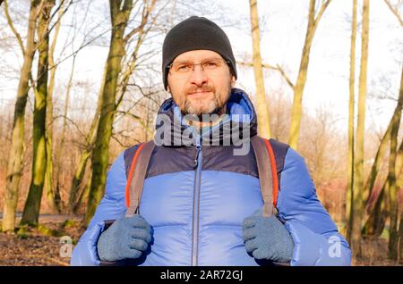 Ein Porträt eines Mannes mittleren Alters in blauer Daunenjacke und ein Rucksack im kargen Wald an einem sonnigen Tag Stockfoto