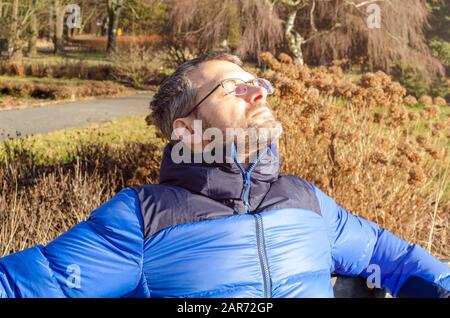 Ein reifer Erwachsener in blauer Daunenjacke sitzt an einem sonnigen Wintertag in der Sonne auf einer Parkbank Stockfoto