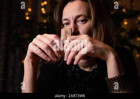Wahrsagerei auf den Runen. Eine reife weibliche Hexe hält eine Rune in der Nähe ihres Gesichts, was "Weltenbaum", "Schutz" bedeutet. Nahaufnahme im Hochformat. Stockfoto