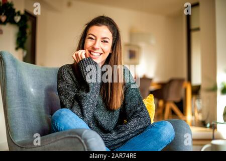 Schuss einer attraktiven lächelnden Frau mit Turtleneck-Pullover und Jeans, während sie sich zuhause im Sessel entspannen. Stockfoto