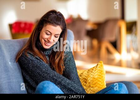 Schuss einer attraktiven lächelnden Frau mit Turtleneck-Pullover und Jeans, während sie sich zuhause im Sessel entspannen. Stockfoto